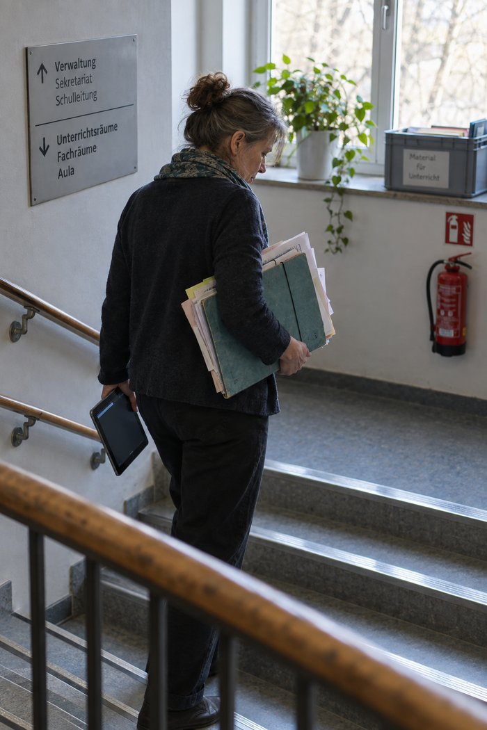 Frau auf dem Treppenabsatz mit einer Sammelmappe unter dem Arm, aus der Blätter in ungleichen Formaten herausragen