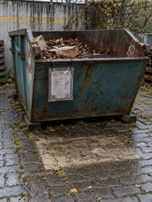 Seit Wochen abgestellte Absetzmulde in der Hofecke mit nassem Laub, ausgeblichenem Auftragszettel in einer Klarsichthülle und dem helleren Abdruck des früheren Standplatzes im Pflaster