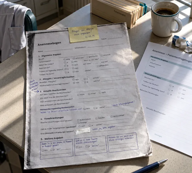 Mehrfach kopierter Anamnesebogen einer Praxis auf einem Tisch im Personalraum, mit handschriftlichen Ergänzungen, einer durchgestrichenen Frage, von Hand gezogenen Kästchen und einem Haftzettel, daneben ein unbeschriebenes Musterformular, eine Kaffeetasse und ein Kasack über der Stuhllehne