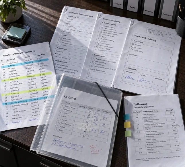 Aufgefächerte Personalformulare eines Klinikträgers auf einer dunklen Holzplatte: Antrag auf Stellenbesetzung, Stellenbeschreibung und Freigabeblatt mit drei Unterschriftsfeldern, daneben die mit Textmarker angestrichene Liste der Gesellschaften und Standorte, ein Tarifauszug mit Haftstreifen und ein Laufzettel im Umlaufdeckel