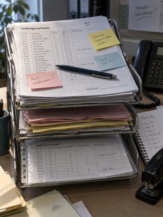 Dreistöckiger Drahtablagekorb in der Verwaltung eines Pflegedienstes zum Monatsabschluss: oben Leistungsnachweise mit leerer Unterschriftenzeile und Haftnotizen zu fehlenden Unterschriften, darunter rosa und gelbe Durchschläge und eine Abgleichliste mit Fragezeichen, daneben Tischtelefon und Rückrufliste