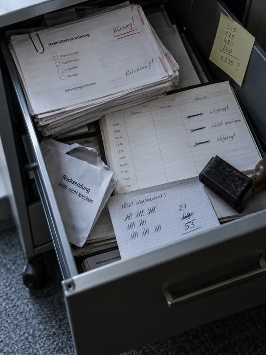 Herausgezogene Schublade eines Büro-Rollcontainers: Heilmittelverordnungen mit roten Kürzungsvermerken, ein Terminbuch mit abgesagten und nicht wahrgenommenen Terminen, eine Strichliste nicht abgerechneter Leistungen, ein aufgerissener Rücksendeumschlag und ein Stempel