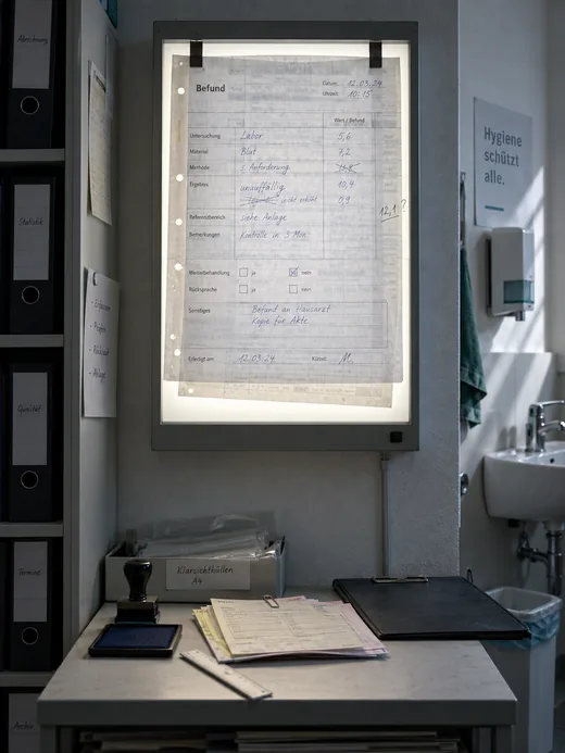 Nebenraum der Verwaltung: Ein handschriftlich ausgefüllter, mehrfach korrigierter Befundbogen hängt am Leuchtkasten, damit die Zahlen überhaupt lesbar werden, darunter auf dem Wandbord Stempel, Lineal und weitere Durchschläge