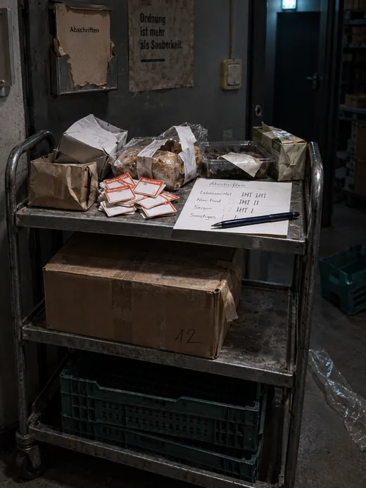 Abschriftenwagen im Nebenlager nach Ladenschluss: oben aussortierte Ware mit eingedrückten Verpackungen, abgezogene Preisetiketten und eine Strichliste mit Kugelschreiber, darunter ein verstaubter Karton, unten leere Steigen, an der Wand ein leerer Halter für den Abschriftenblock