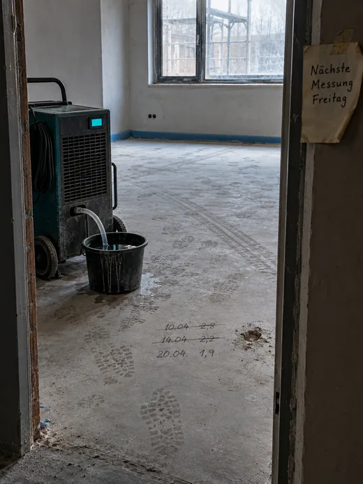 Blick durch die Tür in einen leeren Raum mit Zementestrich voller Schuhabdrücke, links ein Bautrockner mit Schlauch im Eimer, auf dem Estrich drei datierte Messwerte, zwei durchgestrichen, am Türstock ein Zettel zur nächsten Messung