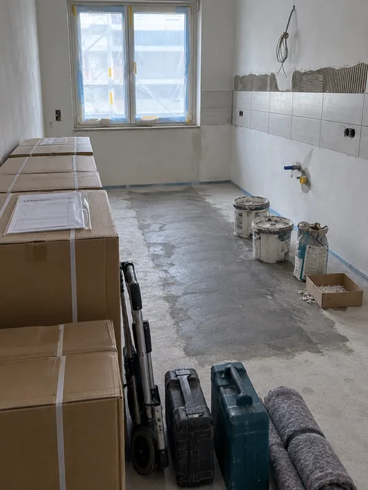 Küchenraum eines Neubaus, der noch nicht bereit ist: feuchter Estrich, halb gesetzter Fliesenspiegel, ein Kabelbündel ohne Dose, davor unangetastet die angelieferten Küchenkartons mit Lieferschein, Sackkarre und Werkzeugkisten