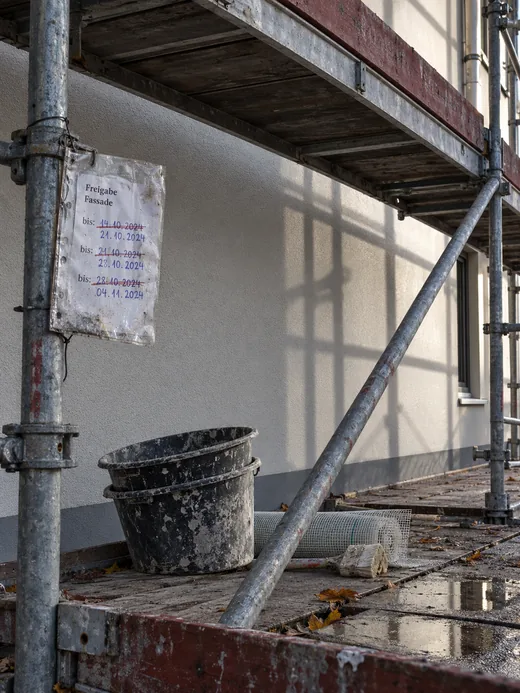 Gerüstfeld vor einer fertig verputzten Fassade, am Stiel eine Freigabekarte mit dreimal verschobenem Datum, auf dem Belag leere Mörtelkübel, ein Rest Armierungsgewebe und eine Regenpfütze