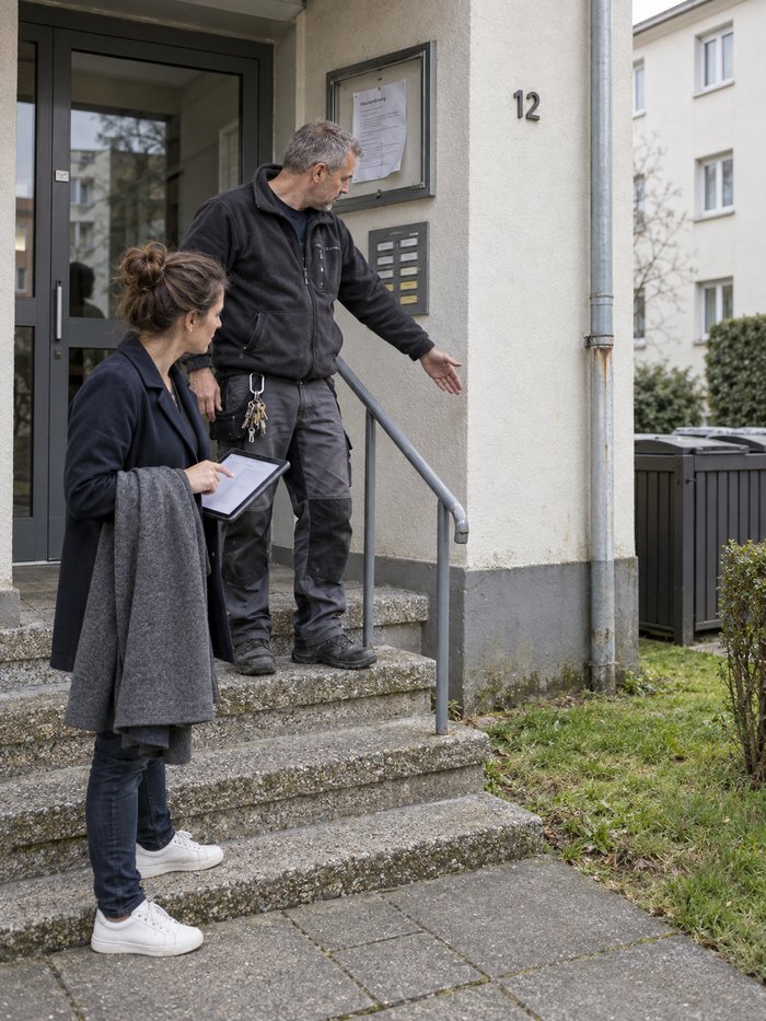 Hauseingang eines Mehrfamilienhauses bei der Begehung: Ein Objektbetreuer in Fleecejacke mit Schlüsselbund am Karabiner steht auf der Waschbetontreppe und deutet an der Fassade entlang, eine Frau mit Mantel über dem Arm und Tablet mit Begehungsliste sieht in dieselbe Richtung
