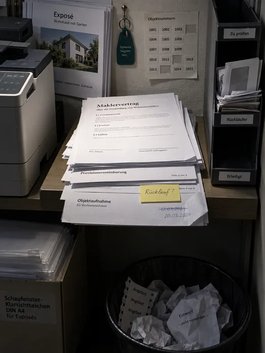 Ablage neben dem Drucker eines Maklerbüros im Licht einer Deckenleuchte: ein Stapel zurückgelaufener Vordrucke, obenauf ein Maklervertrag mit leerem Unterschriftsfeld, darunter eine Provisionsvereinbarung mit fehlender Seite, ein Haftzettel mit der Frage nach dem Rücklauf und eine Objektaufnahme mit überschriebenem Datum, rechts ein Sortierkasten mit überquellendem Fach für Rückläufer, darunter ein Papierkorb mit Fehldrucken