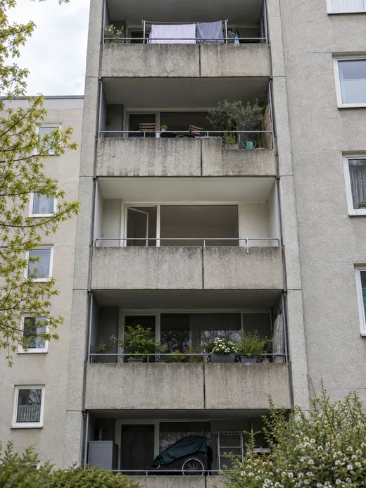 Rückseite einer schlichten Wohnanlage mit fünf Balkonen übereinander: Auf vieren stehen Wäscheständer, Stühle, Kübelpflanzen und ein abgedeckter Fahrradanhänger, der mittlere ist völlig leer, dahinter ein Zimmer ohne einen einzigen Gegenstand