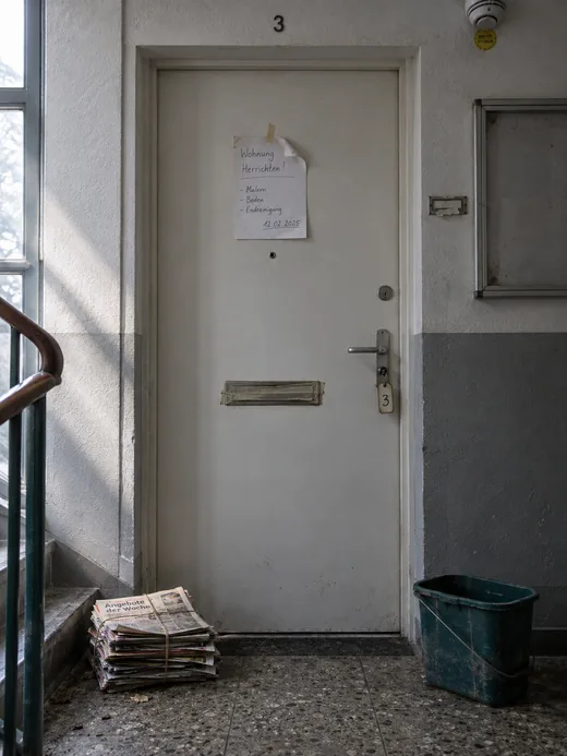 Wohnungstür einer freigezogenen Einheit im Treppenhaus: Auf Augenhöhe ein handgeschriebener Zettel zur Herrichtung mit Malern, Boden und Endreinigung samt Datum, der Briefschlitz mit Klebeband verschlossen, am Drücker ein Schlüsselanhänger mit der Nummer 3, davor ein verschnürter Stapel Wochenblätter und ein abgestellter Eimer, oben ein Rauchmelder mit Prüfplakette und ein leerer Aushangkasten