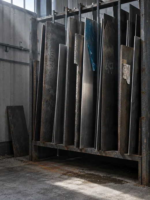 Restblechfach an der Hallenwand: in einem Ständer aus verschweißtem Winkelstahl stehen hochkant angeschnittene Tafeln aus zunderigem Walzstahl, blankem Feinblech und Edelstahl mit halb abgezogener blauer Schutzfolie, auf einer ein eingeschlagener Chargenstempel, auf einer anderen eine verwischte Kreidezahl, an einer dritten ein abgerissener Etikettrest, davor eine an die Wand gelehnte Resttafel