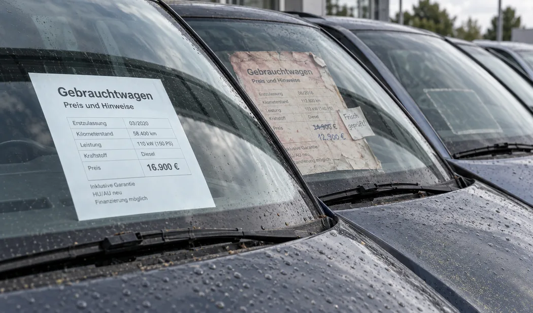 Reihe gebrauchter Fahrzeuge auf dem Freigelände, flacher Blick entlang der verregneten Windschutzscheiben: hinter der ersten Scheibe ein frisch gedrucktes Schild Gebrauchtwagen mit Erstzulassung, Kilometerstand, Leistung, Kraftstoff und 16.900 €, hinter der zweiten dasselbe Schild sonnenverblichen und eingerissen, der Preis von Hand von 14.900 € auf 12.900 € korrigiert und ein halb abgelöster Zettel „Frisch geprüft!“, hinter der dritten Scheibe nur noch der helle Abdruck eines Schildes