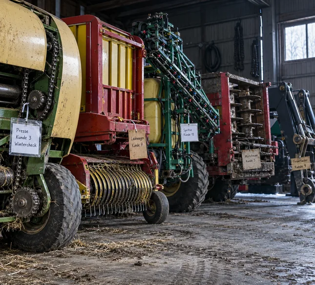 Einlagerungshalle im Winter: Fünf ganz verschiedene Kundenmaschinen stehen nebeneinander, eine Rundballenpresse mit Kettentrieb, ein Ladewagen mit Pick-up, eine Feldspritze mit gefaltetem Gestänge und Düsenreihe, ein Miststreuer mit stehenden Streuwalzen und ein Frontlader, an jeder hängt ein anderer selbst gemachter Anhänger wie „Presse Kunde 3 Wintercheck“ oder „Spritze Kunde 12“, an der Wand Ketten und ein aufgerollter Schlauch