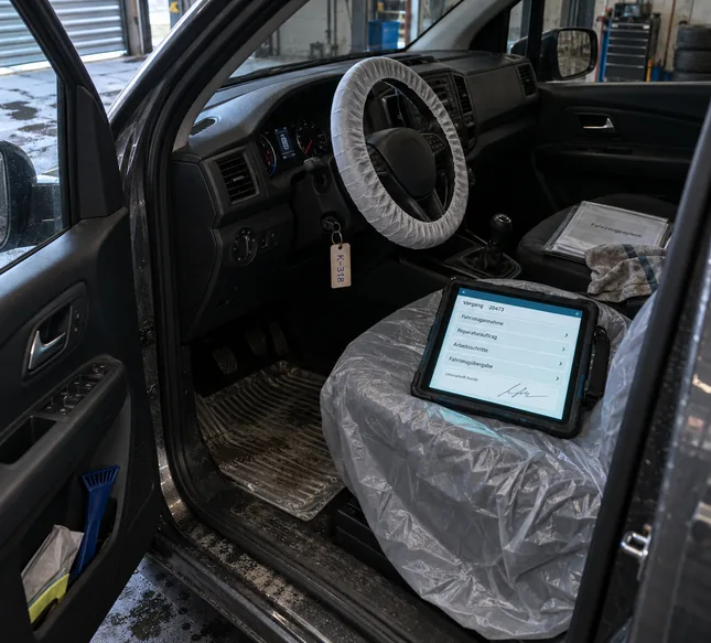 Blick durch die geöffnete Fahrertür in den Innenraum eines Kundenfahrzeugs in der Werkstatthalle: Auf dem mit Schutzfolie bezogenen Fahrersitz liegt ein Tablet in Gummihülle mit Handschlaufe, auf dem Display ein Vorgang mit Fahrzeugannahme, Reparaturauftrag, Arbeitsschritten, Fahrzeugübergabe und der Unterschrift des Kunden, das Lenkrad trägt einen Schonbezug, im Zündschloss steckt ein Schlüssel mit Papieranhänger, auf dem Beifahrersitz liegen eine Mappe mit Fahrzeugpapieren und ein Werkstatttuch, in der Türablage steckt ein Eiskratzer
