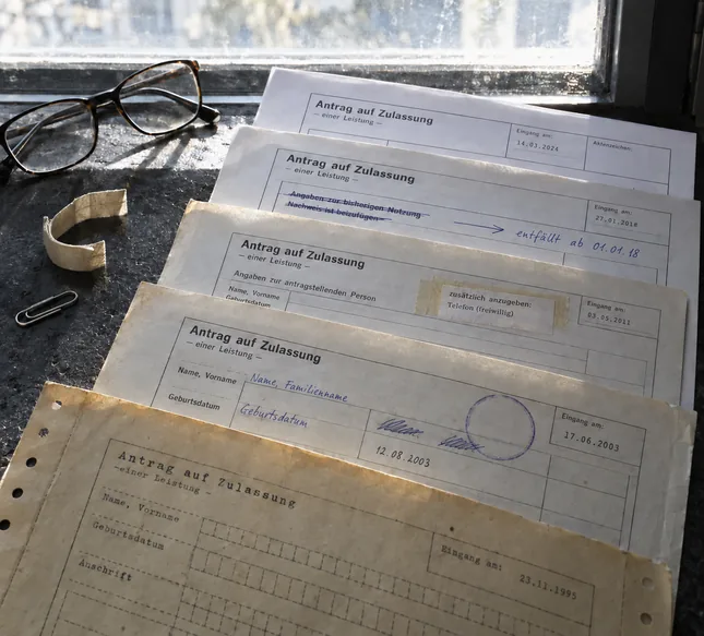 Fensterbank im Gegenlicht: Fünf Fassungen desselben Antragsvordrucks liegen gefächert übereinander, vorn ein vergilbter Bogen im Schreibmaschinenraster von 1995, dann einer mit handschriftlich korrigierten Feldnamen und blassem Stempelkreis, einer mit aufgeklebtem Zusatzfeld, einer mit zwei gestrichenen Zeilen und dem Randvermerk „entfällt ab 01.01.18“ und zuhinterst eine frische Fassung von 2024, daneben eine Lesebrille