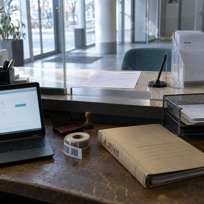 Tresen im Bürgerbüro vor der Öffnung, von der Mitarbeiterseite gesehen: Vorn liegt neben Notebook, Stempelkissen und Etikettenrolle eine Akte, auf deren Deckel von Hand A 27/418 steht, hinter der Acrylscheibe mit Durchreiche ein ausgefülltes Antragsblatt mit derselben Nummer, daneben ein Kugelschreiber an der Kette, ein Fach mit leeren Vordrucken und ein blaugrüner Besucherstuhl