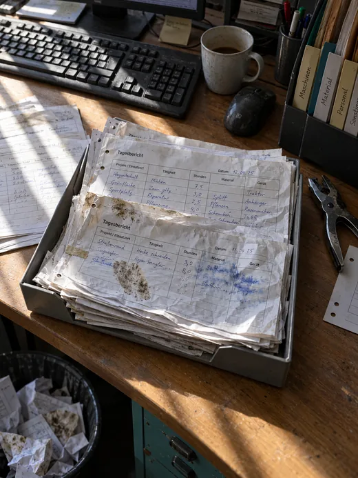 Schreibtischecke im Bauhofbüro im Streiflicht durch Lamellen: In einer Ablageschale aus Metall stapeln sich zerknitterte, verschmutzte Vordrucke der Kolonnen mit handschriftlichen Einträgen zu Einsatzort, Tätigkeit, Stunden, Material und Gerät, auf einem ein erdiger Daumenabdruck und verlaufene Tinte, dahinter Tastatur, Maus und eine Tasse Kaffee, rechts ein Stehsammler mit Registern, unten ein Papierkorb mit zerknüllten Zetteln