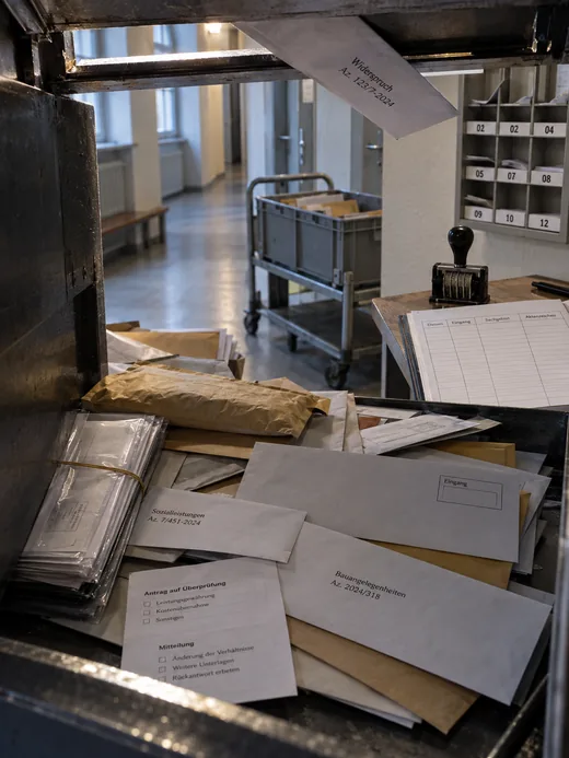 Blick in den geöffneten Sammelkasten eines Nachtbriefkastens im Verwaltungsgebäude vor Dienstbeginn: Umschläge in allen Formaten, ein Versandbeutel, gebündelte Klarsichthüllen und ein loser Vordruck liegen durcheinander, beschriftet mit Sachgebiet und Aktenzeichen, eine Sendung klemmt schräg in der Einwurfklappe, dahinter Datumsstempel und aufgeschlagenes Posteingangsbuch, ein nummeriertes Fächerregal und im Flur ein Rollwagen mit Postkiste