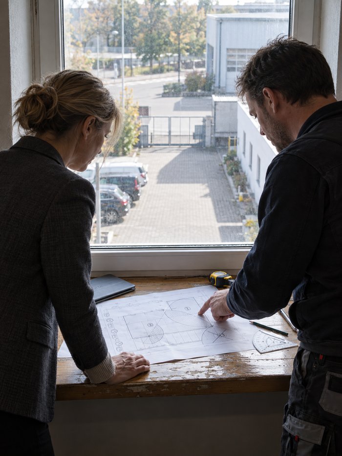 Fensternische im Obergeschoss eines Betriebsgebäudes mit Blick auf Hof, Stellplätze und Toranlage: Ein Techniker in dunkler Arbeitsjacke zeigt auf einen ausgedruckten Lageplan mit eingezeichneten, teils schraffierten Blickfeldern von Kameras, eine Frau in dunklem Blazer stützt sich auf die Fensterbank und sieht auf dieselbe Stelle, daneben ein zugeklapptes Tablet, ein Maßband, ein Bleistift und ein Winkelmesser