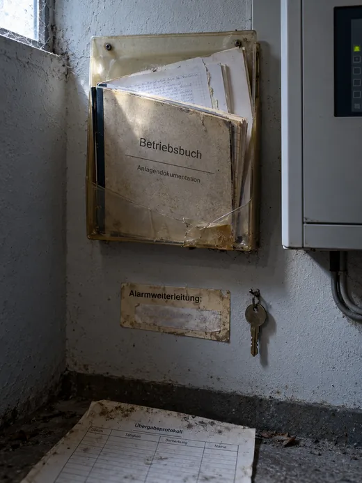 Technikvorraum eines Objekts im Streiflicht: In einem vergilbten, eingerissenen Klarsichthalter an der Wand steckt ein abgegriffenes Betriebsbuch mit losen, handbeschriebenen Blättern, darunter ein verschmutztes Schild Alarmweiterleitung mit überklebter, unleserlicher Nummer und ein einzelner Schlüssel an einem Haken, rechts angeschnitten das Gehäuse einer Zentrale, auf dem Boden ein herausgefallenes, verschmutztes Übergabeprotokoll mit leerer Tabelle
