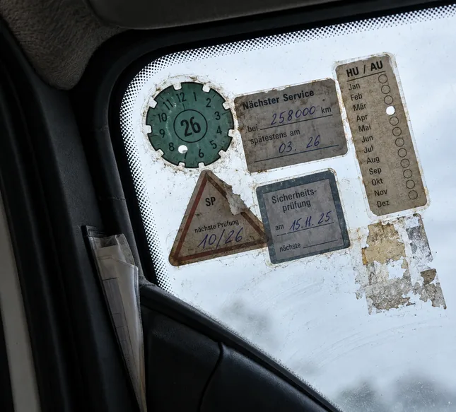 Blick vom Fahrersitz schräg nach oben in die linke obere Ecke einer Windschutzscheibe: Dicht gedrängt kleben dort eine runde grüne Prüfplakette mit der Jahreszahl 26 und gelochtem Monat, ein Werkstattaufkleber Nächster Service bei 258000 km, spätestens 03.26, ein Streifen HU/AU mit den Monaten Januar bis Dezember und einem gelochten Feld, ein dreieckiger Aufkleber SP mit nächster Prüfung 10/26 und abgerissener Ecke, ein Aufkleber Sicherheitsprüfung am 15.11.25 und die abgekratzten Reste eines weiteren Aufklebers, am Scheibenrahmen klemmt eine Klarsichthülle mit einem gefalteten Blatt, durch das Glas fällt flaches graues Tageslicht