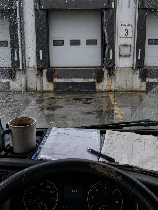 Blick vom Fahrersitz über das abgegriffene Lenkrad durch die regennasse Windschutzscheibe eines stehenden Lkw auf das geschlossene Rampentor eines fremden Betriebs mit ausgerissener Torabdichtung, daneben das Schild Anmeldung 3 und ein leerer, verrosteter Papierhalter, auf dem Beton Pfützen und ein gelber Streifen, auf dem Armaturenbrett ein kalter Kaffeebecher mit eingetrocknetem Rand, ein Frachtbrief in der Klarsichthülle, ein Kugelschreiber und ein Wochen-Tourenplan mit leeren Spalten für Beginn und Ende der Beladung, im Kombiinstrument die Uhrzeit 11:27
