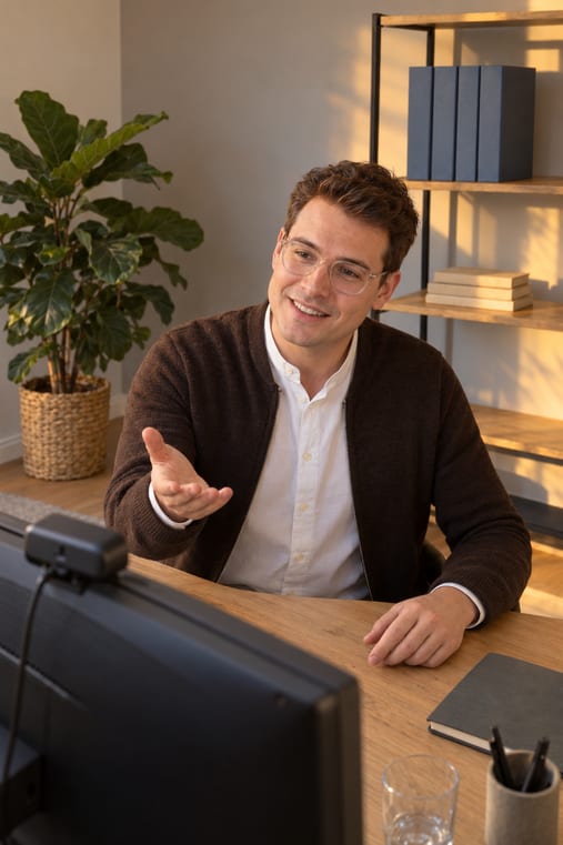 Formilo-Berater mit transparenter Brille und brauner Strickjacke erklärt im Online-Meeting vor seinem Monitor mit Webcam, wie die individuelle Arbeitsauftrag-Vorlage entsteht