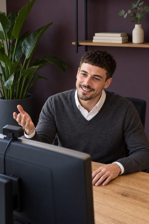 Formilo-Berater mit dunkelbraunem Haar und kurzem Bart im grauen V-Pullover über weißem Hemd erklärt im Online-Meeting vor seinem Monitor mit Webcam mit offen erhobener Hand, wie der individuelle Arbeitsbericht für Ihre Monteure entsteht, hinter ihm eine Strelitzie und ein Wandregal mit Büchern und Eukalyptuszweigen vor pflaumenvioletter Wand