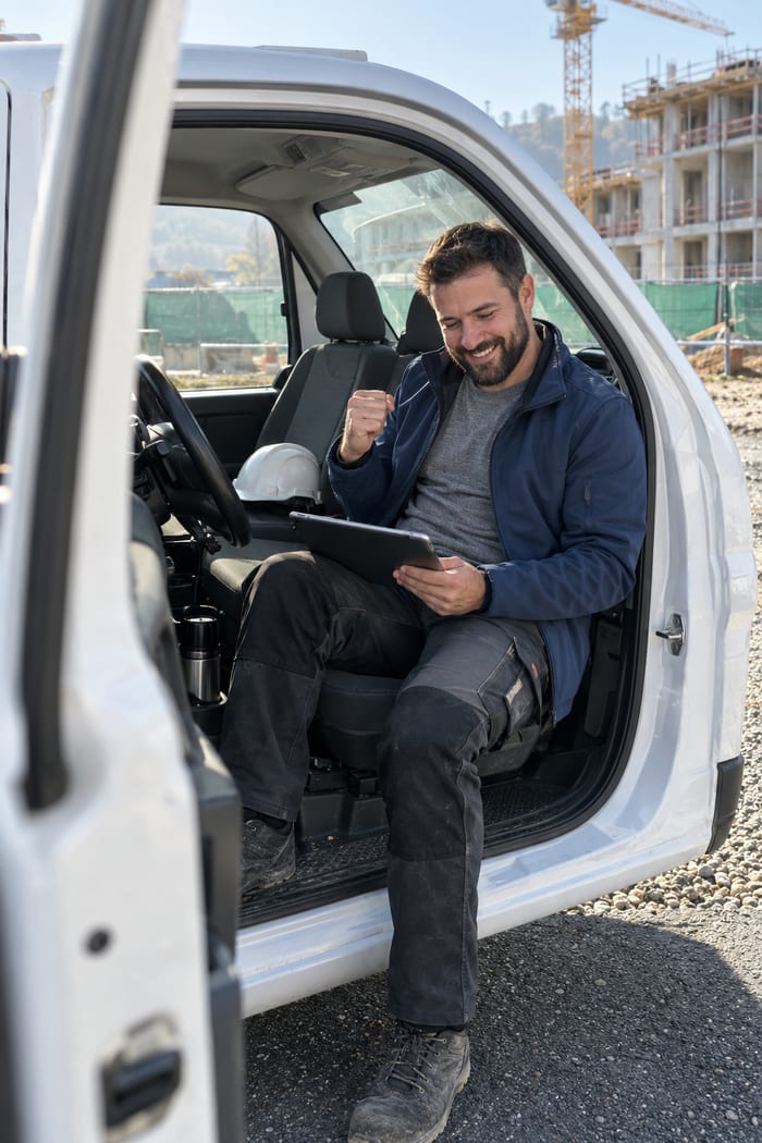 Bauleiter mit dunklem Vollbart sitzt in der offenen Tür seines weißen Transporters vor einer Baustelle mit Kran, lächelt und ballt mit dem Tablet auf dem Knie die Faust