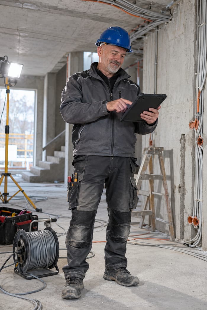 Elektroinstallateurmeister mit blauem Helm im Rohbaugeschoss zwischen Leerrohren, Unterputzdosen und Kabeltrommel tippt zufrieden auf sein Tablet