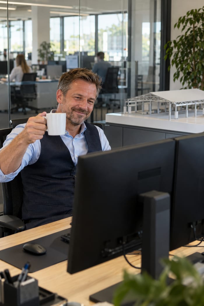 Geschäftsführer eines Hallenbau-Unternehmens hebt im Chefbüro die Kaffeetasse anerkennend in Richtung seiner zwei Monitore, auf dem Sideboard das Modell einer Gewerbehalle