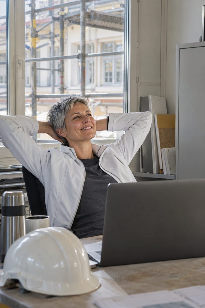 Bauleiterin lehnt sich im Baubüro nach der Bedenkenanmeldung nach VOB am Laptop zufrieden zurück, vor dem Fenster steht das Gerüst an der Fassade
