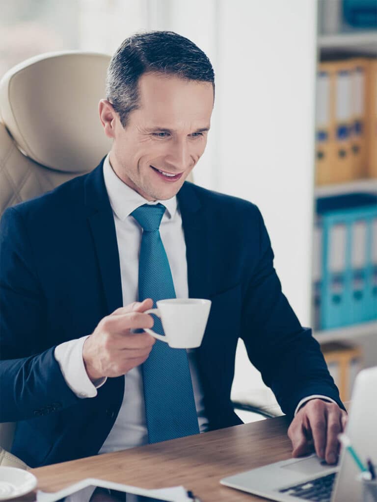Mann im Anzug sitzt mit einem Kaffe am Laptop und lächelt