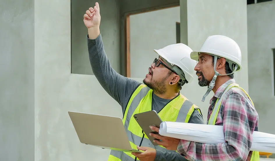 Bauingenieure mit Schutzhelmen und Warnwesten analysieren Strukturelemente eines unvollendeten Wohnbauprojekts.