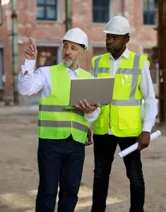 Bauarbeiter mit Laptop erklärt einem Architekten auf der Baustelle alle Besonderheiten des Projekts.
