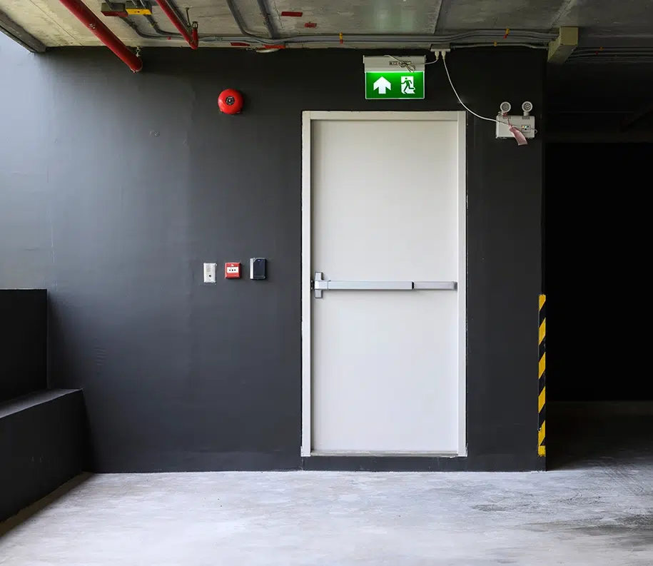 Ein Notausgang mit Schild und roter Alarmglocke über der weißen Tür am Parkhaus.