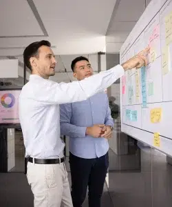 Zwei Projektmanager planen Aufgaben auf einem Kanban-Scrum-Board mit Haftnotizen, zeigen auf den Aufkleber und diskutieren Planung, Strategie und Lösungen unter Verwendung agiler Methoden.