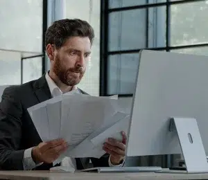 Verärgerter Geschäftsmann sitz am PC und durschaut lauter Papierkram in der Hand