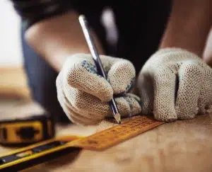 Hände eines Handwerkers in Schutzhandschuhen, der mit Lineal und Bleistift ein Holzbrett misst.