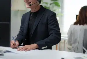 Lächelnder Mitarbeiter mit Headset beim Notizenschreiben während eines Kundensupport-Anrufs