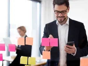 Mann organisiert Aufgaben mit bunten Haftnotizen auf einer Glastafel und hält Smartphone in der Hand.